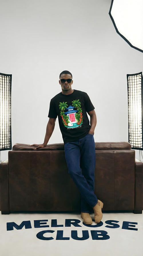 Man wearing a black t-shirt with a colorful design, sitting on a brown couch in a room with 'Melrose Club' branding.